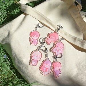 May include: Six pink and white cartoon character keychains with silver chains. The keychains are on a white canvas tote bag.
