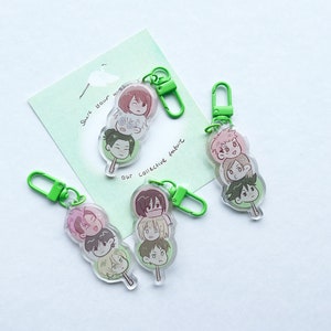 Puede incluir: Tres llaveros verdes con dijes de acrílico transparente que representan personajes de dibujos animados apilados uno encima del otro. Los dijes tienen forma de dango, un dulce japonés. El texto en la tarjeta dice "Comparte tu mundo, nuestra cultura colectiva."