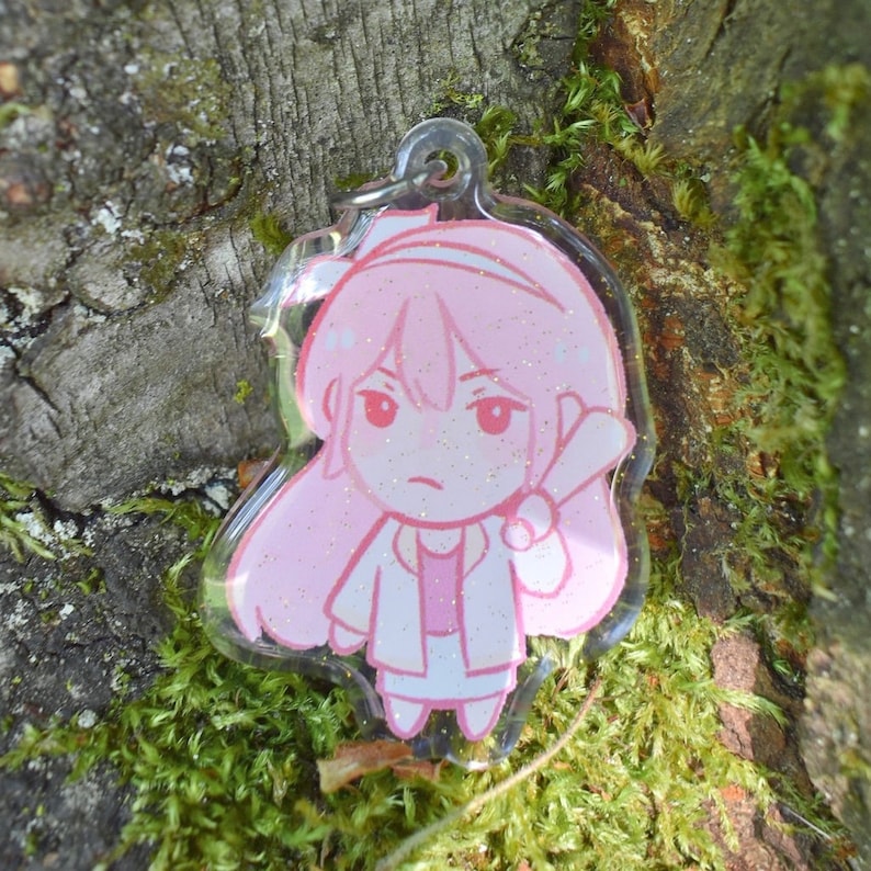 May include: Pink glitter keychain featuring a cartoon character with long pink hair, wearing a white jacket and a pink shirt. The character is holding a white object in their right hand.