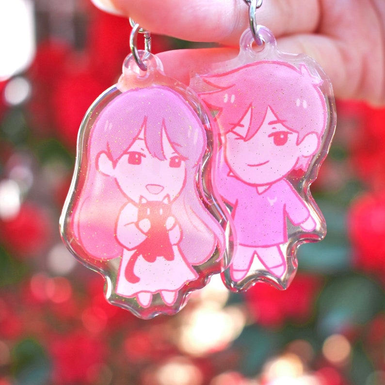 May include: Two pink glitter charm keychains featuring cartoon characters. The female character is holding a black cat. The male character is wearing a pink shirt and has pink hair.