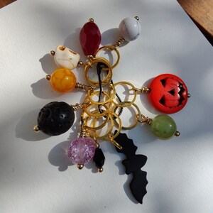 Op de afbeelding: Een verzameling Halloween-thema bedels, waaronder een uitgesneden oranje pompoen, een zwarte vleermuis en diverse kralen in kleuren als rood, wit, geel en paars. De bedels zijn verbonden met goudkleurige ringen.