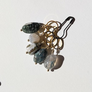 Op de afbeelding: Een verzameling stekenmarkeerders van edelstenen. De markeerders hebben gepolijste, ovaalvormige stenen in tinten groen, wit en blauw. Elke steen is bevestigd aan een goudkleurige ring met een kleine gouden kraal. Een zwarte veiligheidsspeld is ook inbegrepen.