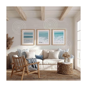 May include: A living room scene featuring three framed ocean wave prints above a white sofa with decorative pillows and a blue throw. A wooden chair and woven side table complete the coastal decor. The room has a neutral color palette.