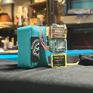 May include: A teal blue zippered pouch with a pink zipper and a black and white logo. Beside it are three stacked chalk cubes with the brand names "Pioneer" and "Tweeten" visible. The items are on a dark surface.
