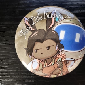 May include: A round button with a cartoon illustration of a brown-haired character with bunny ears, wearing a brown jacket and holding a rolled-up scroll. The character is looking to the right with a serious expression. The background is a light brown color with the text "Fussy Little Bunbun B" in white. The bottom right corner of the button has the text "@aikur Arts" in white.