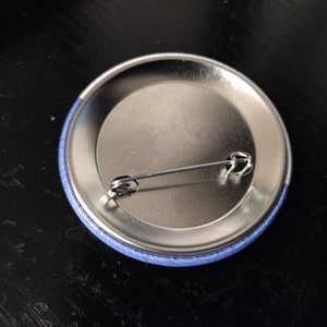 May include: The back of a blue button with a silver pin attached.
