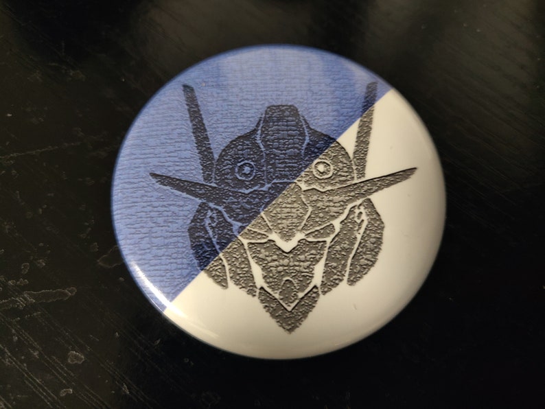 May include: A white and blue button with a black and white graphic of a robot head. The robot head is stylized and has a textured look.