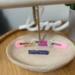 May include: Two hair clips, one pink and white with glitter, and one blue and purple with glitter, on a beige textured surface.