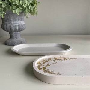 May include: Two white oval-shaped concrete trays. One tray has a gold leaf design. Both trays are on a white surface.