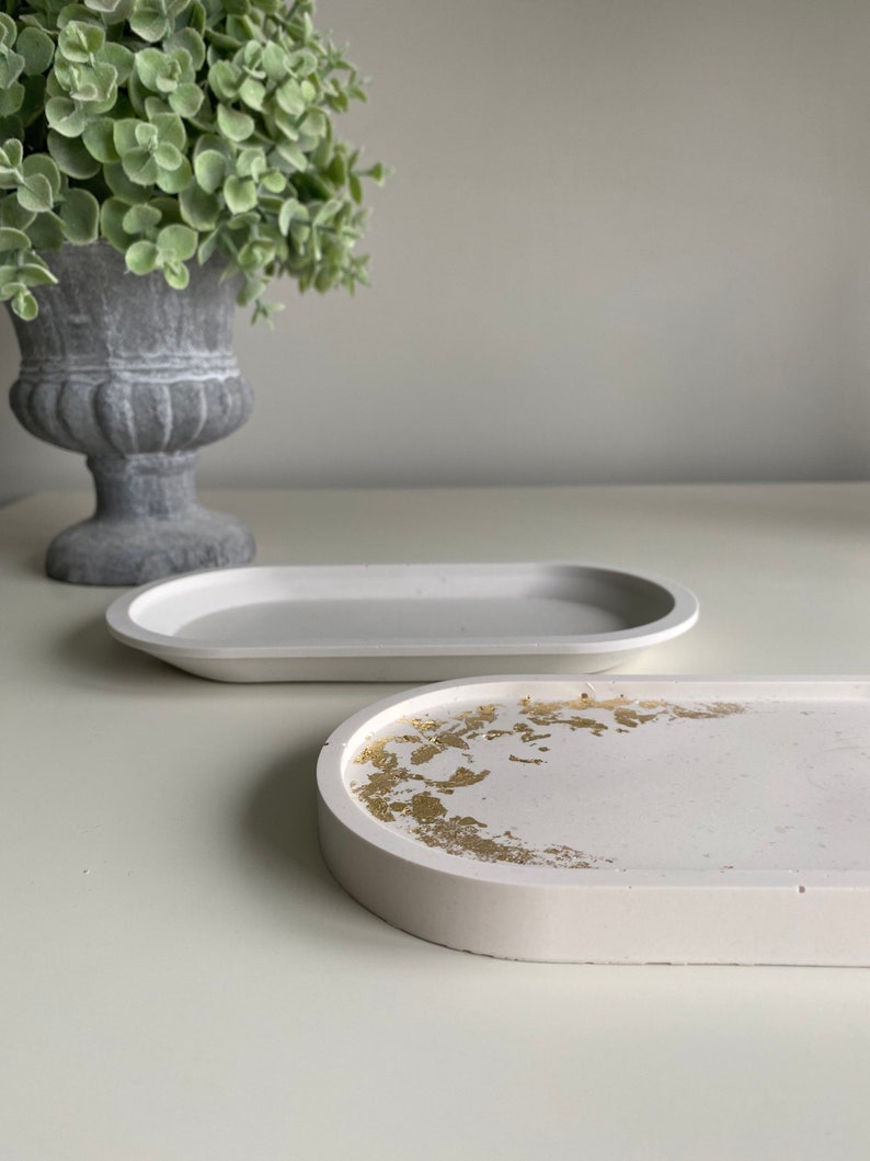 May include: Two white oval-shaped concrete trays. One tray has a gold leaf design. Both trays are on a white surface.