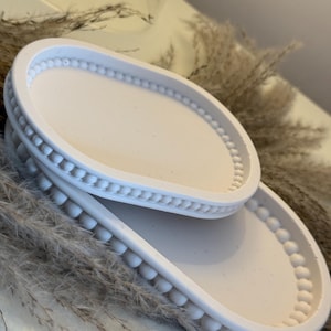 May include: Two white, oval trays are stacked, featuring a minimalist design. The trays have a beaded border and a smooth interior. The background includes dried ornamental grass, adding natural texture and neutral tones.