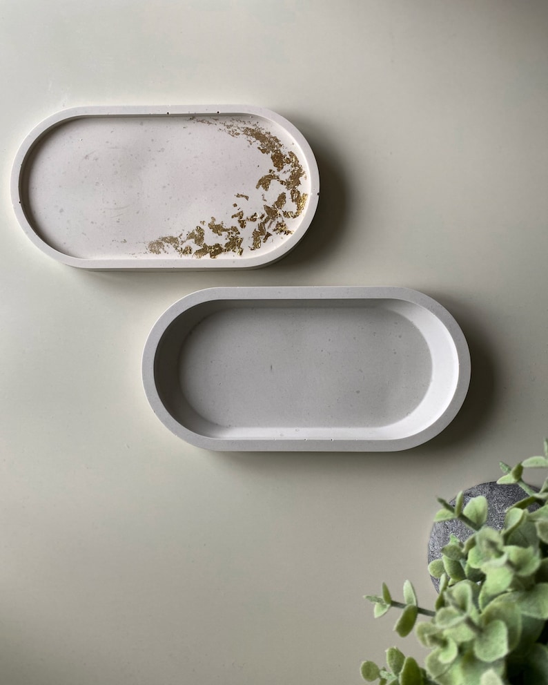 May include: Two oval-shaped concrete trays. One tray has a white background with gold leaf accents. The other tray is plain white.