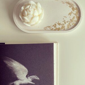 May include: A white oval concrete tray with gold leaf accents. A white flower-shaped candle sits on the tray.