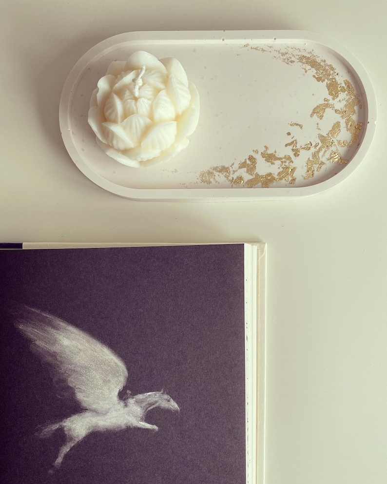 May include: A white oval concrete tray with gold leaf accents. A white flower-shaped candle sits on the tray.