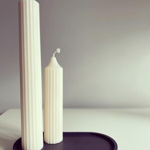 May include: Two white pillar candles of different sizes on a black oval tray. The candles have a ribbed texture.