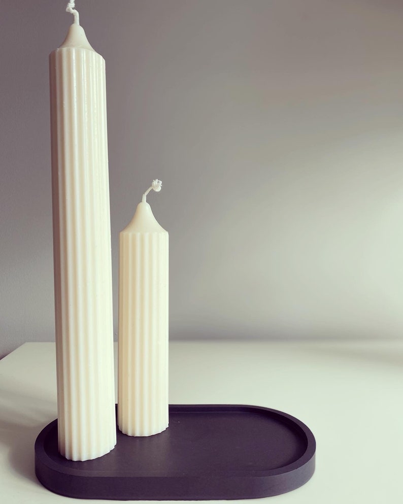 May include: Two white pillar candles of different sizes on a black oval tray. The candles have a ribbed texture.
