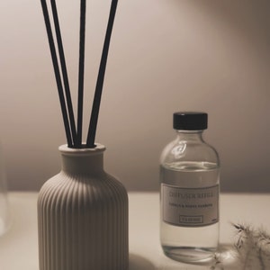 May include: A white ceramic diffuser bottle with black reeds and a clear glass bottle with a black cap and a label that reads "Diffuser Refill Citrus & White Verbena VK Home".