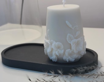 Puede incluir: Vela blanca con un diseño floral en una bandeja ovalada negra. La vela tiene una mecha blanca y está rodeada de flores blancas.