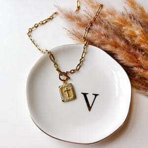 May include: A gold-colored necklace with a rectangular pendant featuring a cross design. The necklace rests on a white plate with a gold rim and the letter "V". Dried ornamental grass is in the background.