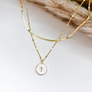 Pode incluir: Um colar dourado em camadas sobre um fundo branco. A camada superior é uma corrente plana, enquanto a segunda apresenta pequenas contas e um pingente com um centro branco e uma cruz dourada. Grama ornamental seca ao fundo.
