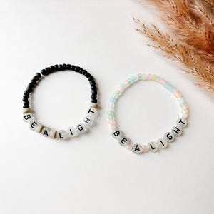 May include: Two beaded bracelets. One is black with white letter beads spelling "BE A LIGHT". The other is pastel colored with the same message. Both bracelets are on a white surface with dried plants in the background.