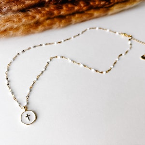 May include: Gold-coloured necklace with a white pendant featuring a black cross. The delicate chain is composed of small, linked segments. The pendant is round with a gold border. The necklace is displayed on a white surface.