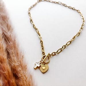 May include: Gold-toned necklace with a toggle clasp and three charms: a cross, a heart, and a sunburst heart. The chain is a chunky link style. The necklace is displayed on a white surface with dried ornamental grass.