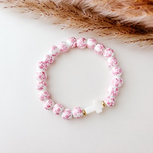 May include: White beaded bracelet with a small white cross charm. The beads are decorated with a pink floral pattern.