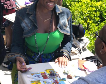 Puede incluir: Una mujer con una camiseta verde y una chaqueta vaquera está sentada en una mesa con cartas del tarot. Se ve un collar con la palabra "intuición". La mesa está cubierta con un paño blanco y hay varias cartas del tarot y cristales.