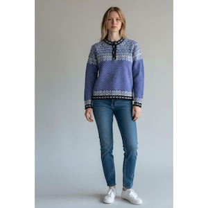 May include: A woman wearing a blue sweater with white geometric patterns and a button-up collar. The sweater is paired with blue jeans and white sneakers. The background is a solid light gray.