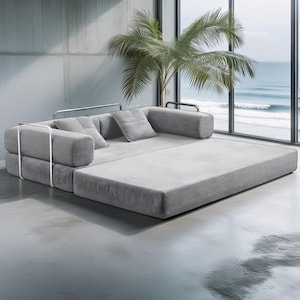 May include: A modern gray sectional sofa with a minimalist design. The sofa features a low profile, a pull-out section, and several matching throw pillows. The frame is accented with silver metal supports. The background includes a large window with a view of the ocean and a palm tree.