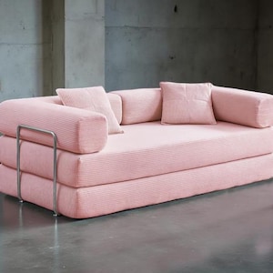 May include: A pink corduroy sofa with a minimalist design. The sofa features a stacked construction with rounded armrests and a metal frame. Includes two square and two cylindrical pillows. The sofa is set against a concrete wall.