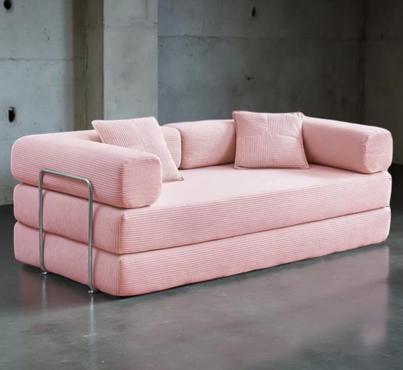 May include: A pink corduroy sofa with a minimalist design. The sofa features a stacked construction with rounded armrests and a metal frame. Includes two square and two cylindrical pillows. The sofa is set against a concrete wall.
