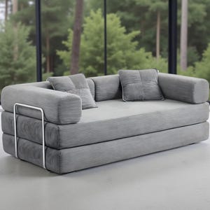 May include: A modern gray sofa with a minimalist design. The sofa features a corduroy-like texture and includes two square throw pillows. The frame has a silver metal accent. The sofa is a stylish addition to any living space.