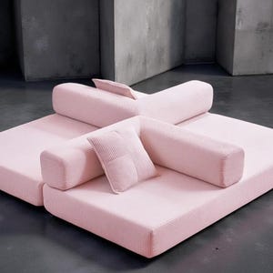 May include: A modular, light pink sofa set with a corduroy texture. The set includes two large square base cushions, cylindrical backrests, and a small square pillow. The design is modern and minimalist, suitable for a living room.