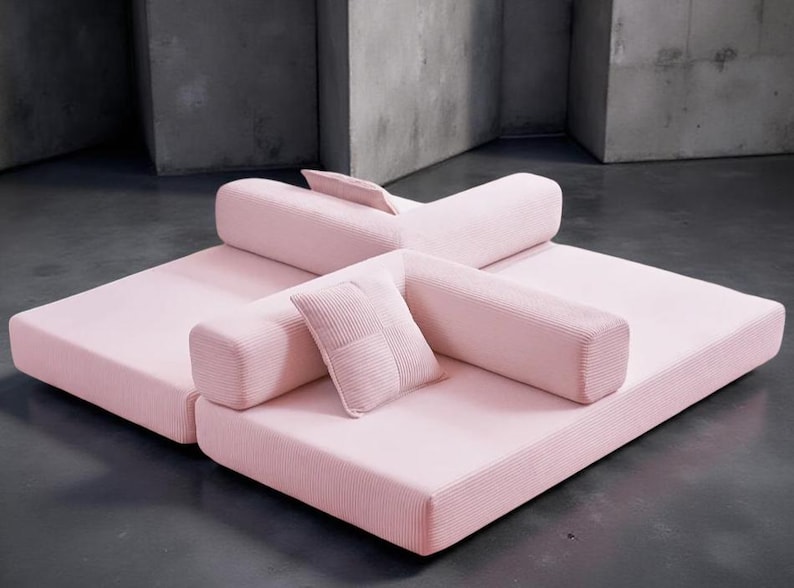 May include: A modular, light pink sofa set with a corduroy texture. The set includes two large square base cushions, cylindrical backrests, and a small square pillow. The design is modern and minimalist, suitable for a living room.
