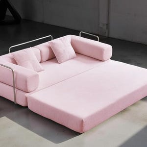 May include: A modern, pink corduroy sofa with a pull-out section. The sofa has a rectangular shape with rounded armrests and two square pillows. The frame is made of silver metal. The sofa is designed for comfort and style.
