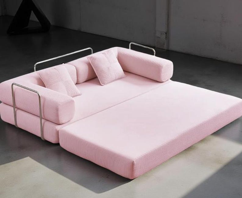 May include: A modern, pink corduroy sofa with a pull-out section. The sofa has a rectangular shape with rounded armrests and two square pillows. The frame is made of silver metal. The sofa is designed for comfort and style.