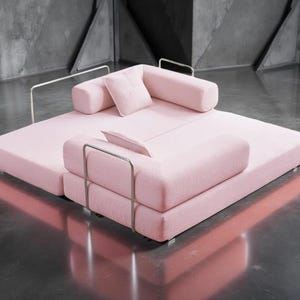 May include: A modern, modular sofa in a soft pink hue. The sofa features a low profile with a rectangular base and rounded bolster cushions. Silver metal armrests add a sleek contrast. Includes a small square pillow.