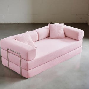 May include: A pink, modular sofa with a minimalist design. The sofa features a low profile, with a stacked base and rounded armrests. Two small square pillows and two bolster pillows are included. A silver metal frame adds a modern touch. The sofa is made of a ribbed fabric.