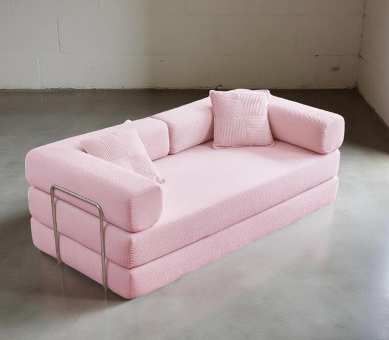 May include: A pink, modular sofa with a minimalist design. The sofa features a low profile, with a stacked base and rounded armrests. Two small square pillows and two bolster pillows are included. A silver metal frame adds a modern touch. The sofa is made of a ribbed fabric.