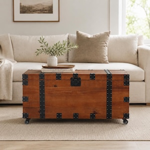 Op de afbeelding: Een rechthoekige houten kist met een warme bruine afwerking, geaccentueerd met zwarte metalen banden en hoekbeugels. De kist heeft een zwarte sluiting en staat op kleine wielen. Hij staat voor een beige bank.