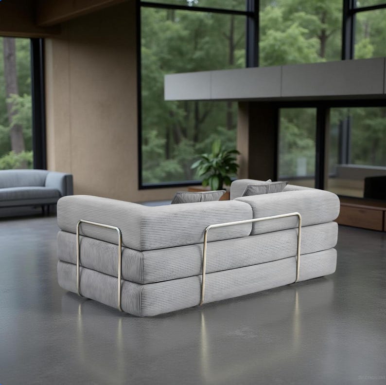 May include: A modern, light gray sectional sofa with a unique stacked design. The sofa features a corduroy-like texture and is supported by polished metal frames. The room has large windows with a view of a forest.