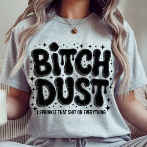 May include: A light gray t-shirt with the words "Bitch Dust" in large, black, bubble letters. Below the text is the phrase "I sprinkle that shit on everything" in a smaller font. The design is surrounded by small star shapes.