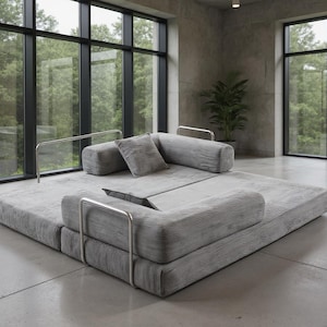May include: A modern gray modular sofa with a low profile and a corduroy texture. The sofa features a square base with attached bolster pillows and a square pillow. Polished metal armrests add a sleek touch. The sofa is in a room with large windows.