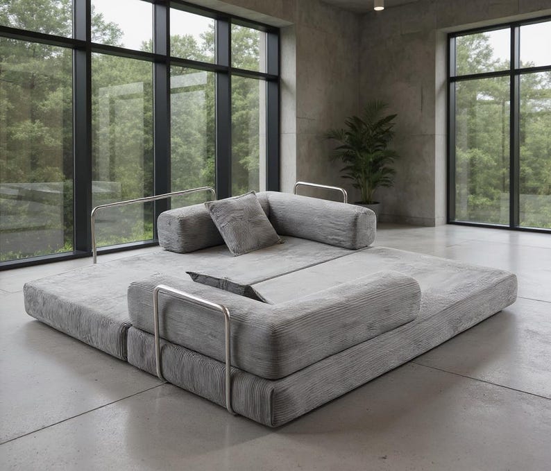 May include: A modern gray modular sofa with a low profile and a corduroy texture. The sofa features a square base with attached bolster pillows and a square pillow. Polished metal armrests add a sleek touch. The sofa is in a room with large windows.
