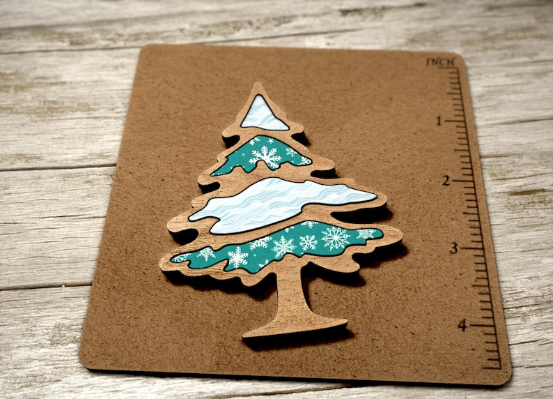 May include: A teal and white wooden Christmas tree ornament with a snowflake pattern. The ornament is approximately 7.5 cm tall and is on a brown background.