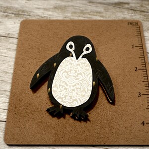 May include: A black and white penguin cutout with a white damask pattern on its belly. The penguin is approximately 7.6 cm tall and is on a brown surface with a ruler in inches.