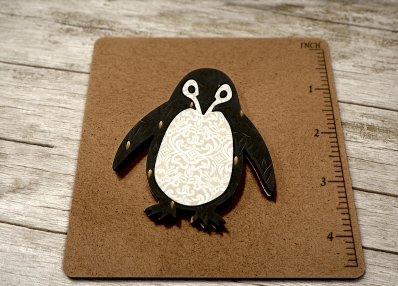 May include: A black and white penguin cutout with a white damask pattern on its belly. The penguin is approximately 7.6 cm tall and is on a brown surface with a ruler in inches.