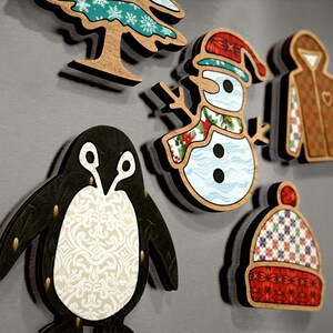 May include: A set of six wooden Christmas decorations featuring a penguin, snowman, winter hat, tree, and two winter jackets. The decorations are decorated with various patterns and colours, including red, green, blue, white, and brown.
